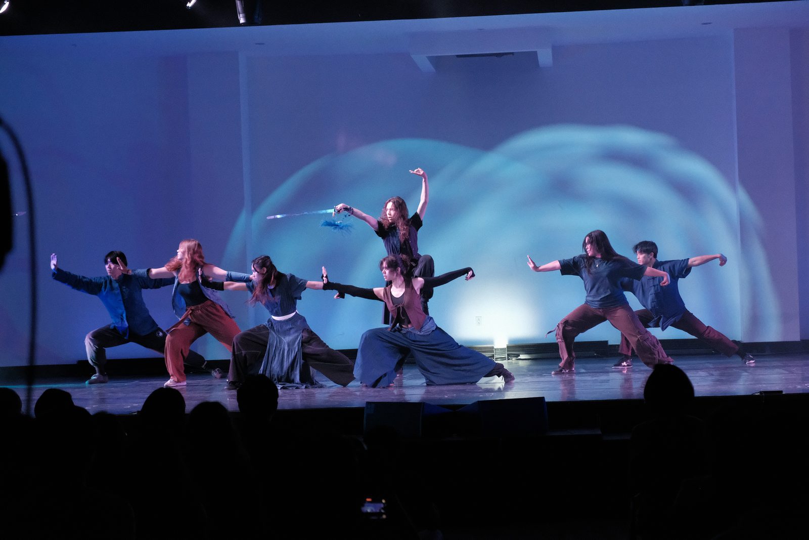 Wushu club members in wide low stances across the stage, lit blue with a swirling projection behind them