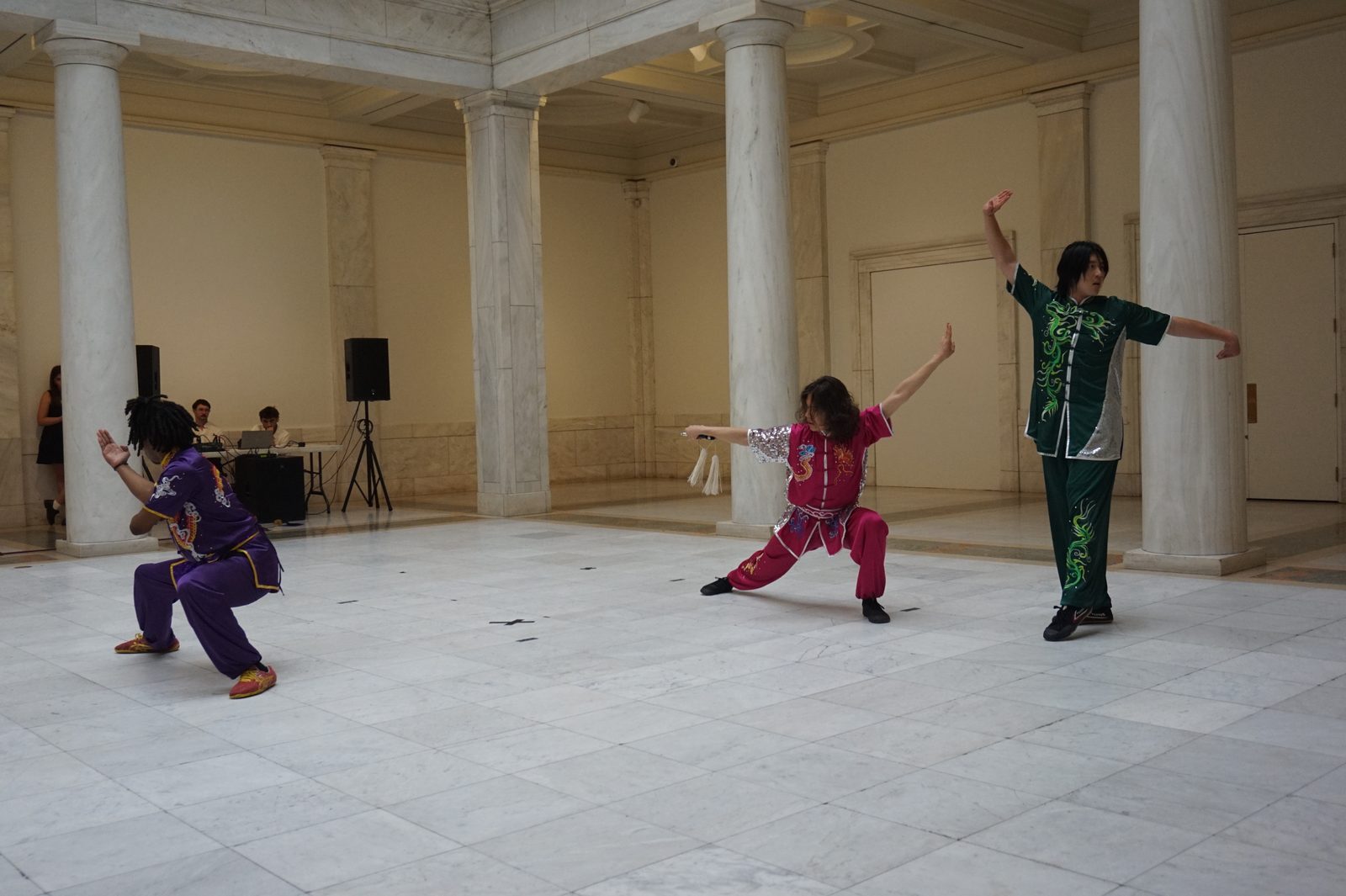 Three performers in coloured uniforms holding stances in the museum's marble columned hall