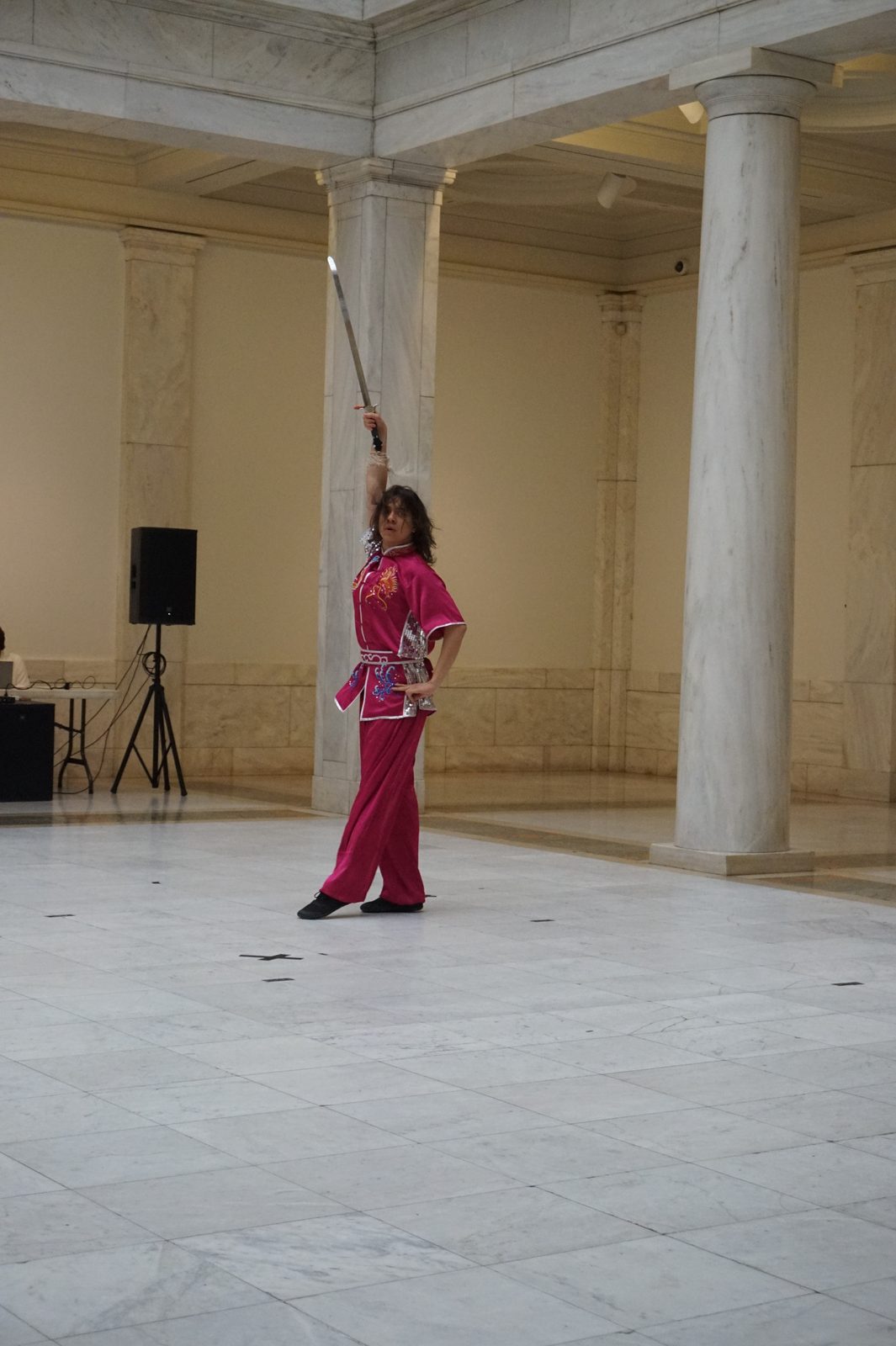 A performer in a pink uniform holding a staff vertically overhead between marble columns